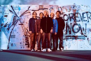 Five individuals stand confidently in front of a wall covered with posters and graffiti, conveying themes of protest and urgency.