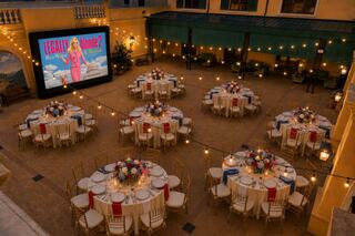 An elegant courtyard features round tables adorned with flowers and candles, illuminated by string lights, and a large screen displaying "Legally Blonde 2."
