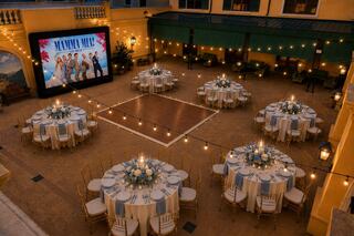 A beautifully arranged outdoor venue with round tables decorated for an event, featuring a large screen displaying "Mamma Mia!" and string lights.