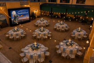 An elegant outdoor setup features round tables adorned with floral centerpieces, strung lights, and a large screen displaying "Ratatouille."