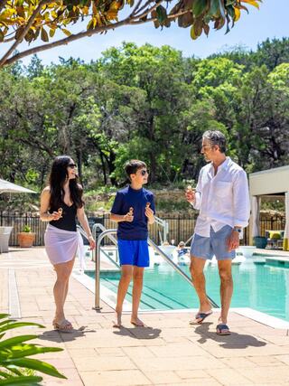 A woman, a boy, and a man in casual summer attire enjoy a sunny day by a pool surrounded by lush greenery.