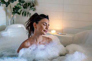 A woman relaxes in a bubble-filled bath, eyes closed, exuding tranquility amidst candlelight and green foliage.