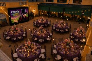 A beautifully decorated courtyard features round tables with floral centerpieces, twinkling lights, and a screen showcasing "Charlie and the Chocolate Factory."