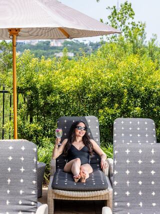 A woman lounges by a pool in a black swimsuit, sipping a colorful drink, surrounded by lush greenery and sun chairs under a parasol.