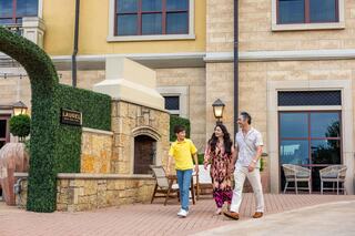 A family strolls together outside a stylish building, enjoying a sunny day. The setting features decorative plants and elegant architecture.