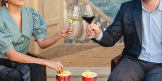 Two people clink glasses of wine and champagne, sharing popcorn in colorful containers, against a backdrop of art and decor.