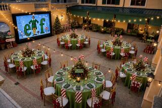 An elegantly decorated outdoor space features festive tables with green and red themes, surrounded by holiday decorations and a screen displaying "Elf."