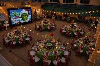 A festive courtyard features beautifully decorated tables with greenery and holiday accents, alongside a large screen playing a Christmas movie.