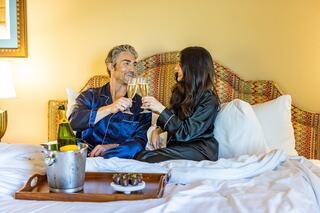 A couple in pajamas cheers with champagne glasses while sitting on a bed, surrounded by a cozy atmosphere and a tray of treats nearby.