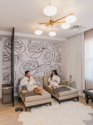 A couple relaxes in a cozy spa-like room, dressed in robes, surrounded by floral decor and soft lighting.