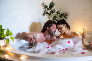 A couple enjoys a romantic bubble bath, sipping drinks amidst candles and rose petals, sharing intimate smiles and laughter.