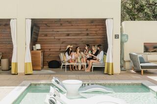 A group of women enjoy drinks and conversation in a cozy, shaded lounge area by a pool, surrounded by modern decor and greenery.