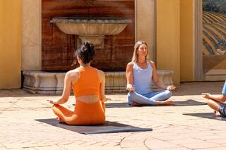 Two women practice yoga outdoors, seated in meditation poses, with a fountain in the background and a serene atmosphere.