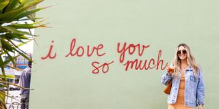 A woman stands smiling in front of a green wall with red graffiti that reads, "i love you so much," surrounded by plants.