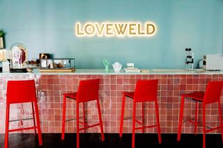 A vibrant café features a pastel blue wall, red stools at a sleek counter, and a glowing "LOVEWELD" sign, creating a welcoming atmosphere.