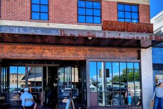 A trendy café with large glass windows and a rust-colored sign, inviting patrons to enjoy a casual atmosphere. Outdoor seating is visible.