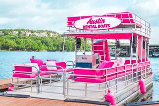 A vibrant pink party boat docked by the water, featuring plush seating and a stylish design, perfect for a fun day on the lake.