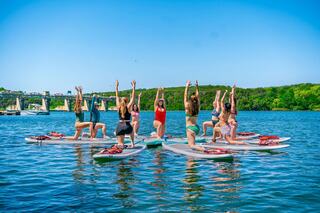 A group of women practice yoga on paddleboards in a sunny lake, showcasing various poses against a backdrop of lush greenery.