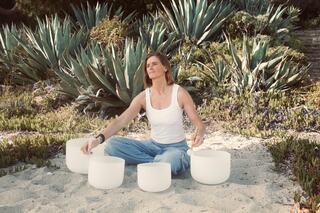 A woman in a white tank top sits on the sand, surrounded by large plants, playing crystal singing bowls with a peaceful expression.