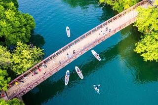 A scenic view of a bridge over turquoise water, with people walking on it and boats paddling below, surrounded by lush greenery.
