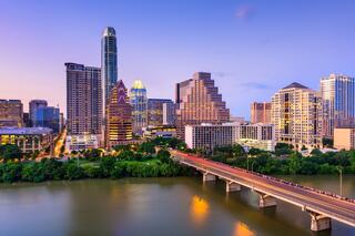 A vibrant city skyline at dusk, featuring a mix of modern skyscrapers, lush greenery, and a busy bridge reflecting the evening light.