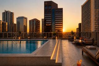 A rooftop pool glistens at sunset, surrounded by modern skyscrapers, with cozy seating and glowing lights creating a tranquil atmosphere.