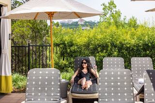 A woman relaxes on a lounge chair by a sunny poolside, sipping a drink under an umbrella, surrounded by lush greenery.