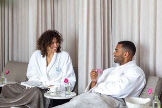 A woman and man in spa robes enjoy a relaxed moment together, smiling and chatting, surrounded by soft décor and light refreshments.