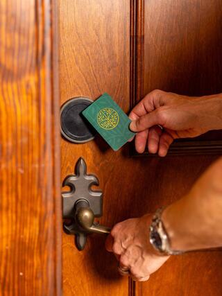 A hand holds a green access card near a door lock, preparing to unlock it while another hand grasps the door handle.