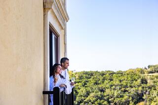 A couple in bathrobes enjoys drinks on a balcony, smiling and looking out at a lush green landscape under a clear blue sky.