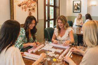A group of women enjoy a lively game of mahjong at a cozy table, laughing and engaging, with drinks and a warm atmosphere.