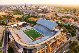 Aerial view of a large stadium surrounded by a vibrant cityscape, showcasing a sports field with green turf and a backdrop of urban buildings.