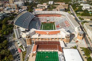 Aerial view of a large sports stadium surrounded by a cityscape, featuring a green football field with "TEXAS" marked in the center.