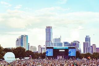 A vibrant festival scene unfolds with a massive crowd gathering in front of a stage, framed by a striking city skyline under a blue sky.