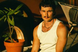 A man with a mustache and a tank top sits outdoors, exuding a relaxed vibe, with greenery and a tire in the background.