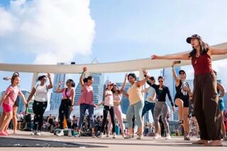 A diverse group of people dances joyfully outdoors, surrounded by urban scenery and a vibrant atmosphere under a bright sky.