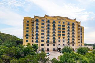 A tall, yellow hotel building stands amidst lush greenery, featuring multiple balconies and a welcoming entrance. Clear skies enhance the scene.