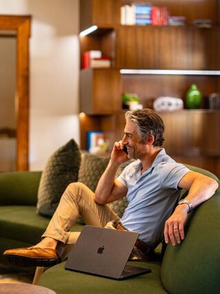 A man sits comfortably on a green couch, talking on the phone, with a laptop beside him and a cozy, modern decor in the background.
