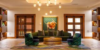 A cozy lobby features a green sofa, cushioned chairs, a round wooden coffee table, and artistic decor, illuminated by stylish pendant lights.