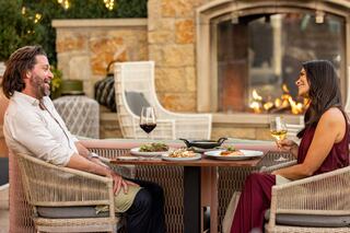 A couple shares a cozy dinner on a patio, enjoying delicious food and drinks, with a warm fireplace setting the mood.