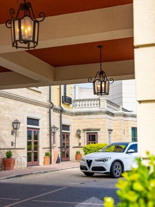 A stylish car is parked beneath decorative lanterns and near elegant architecture, surrounded by greenery and pots.