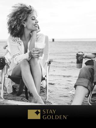 A smiling woman with curly hair enjoys a drink while sitting on the beach, sharing a moment with a companion. The vibe is relaxed and cheerful.