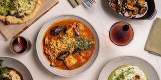 A beautifully arranged table featuring pasta with mussels in a rich sauce, a side of seafood, risotto, and a glass of dark beverage.