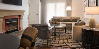 A cozy, modern hotel suite living space featuring a comfortable sofa, fireplace, dining area, stylish decor, and a well-equipped kitchen. Warm lighting enhances the ambiance.