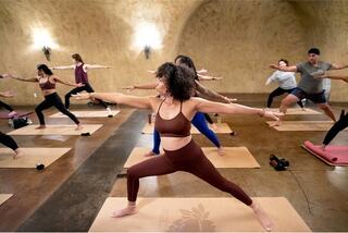 A woman wearing a brown fitness outfit, standing on a yoga mat in warrior two pose in a group yoga class