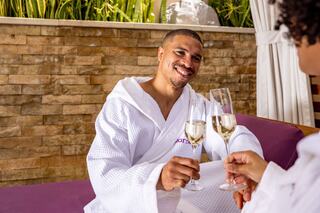 A smiling man and woman in spa robes toast with champagne glasses, enjoying a luxurious and relaxing moment together.