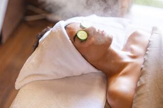 A person relaxes on a spa table, covered with a towel and topped with cucumber slices, enjoying a soothing steam treatment.