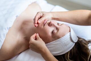 A woman relaxes during a facial treatment, her eyes closed and a headband on, as a therapist gently works on her face.