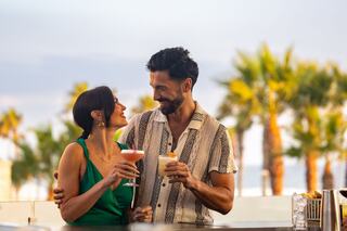 A couple shares a joyful moment at a bar, holding cocktails and smiling, with palm trees and a vibrant sky in the background.