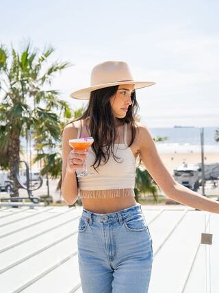 A woman in a crop top and wide-brimmed hat holds a colorful drink, with a sunny beach and palm trees in the background.